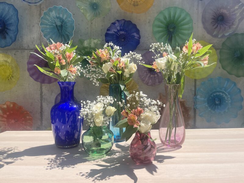 Image shows a variety of vases with bouquets in them in front of a colorful glass wall.