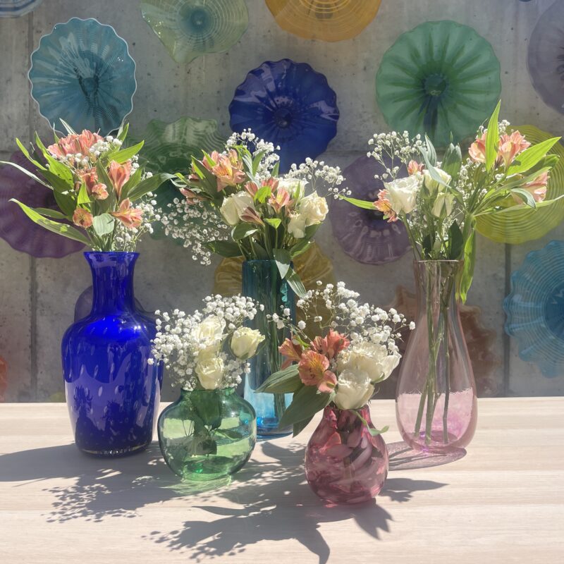 Image shows a variety of vases with bouquets in them in front of a colorful glass wall.