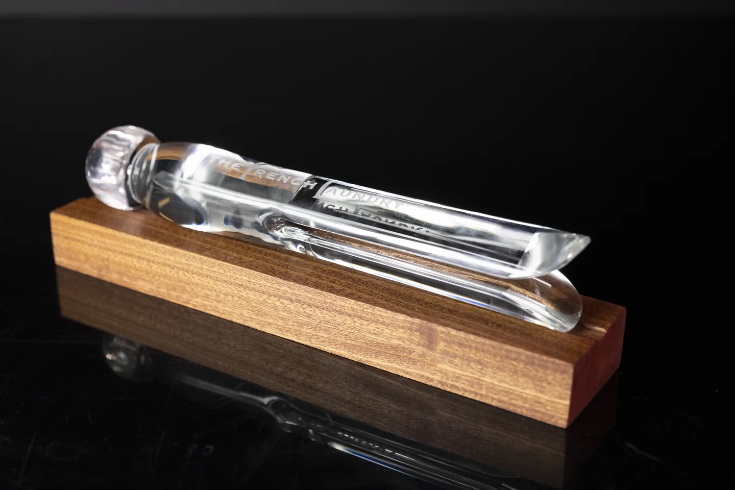 Clear glass pin-shaped piece with an inscription, resting on a wooden base.